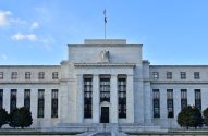 Edifício Federal Reserve: fachada de mármore branco, com águia, colunas e bandeiras sob céu azul.