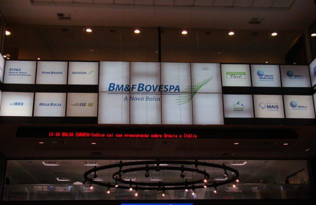 Painel de bolsa de valores com logos BM&FBOVESPA e ticker: "BOLSA EUROPA-Indice cai con preocupação sobre Grécia e Itália".