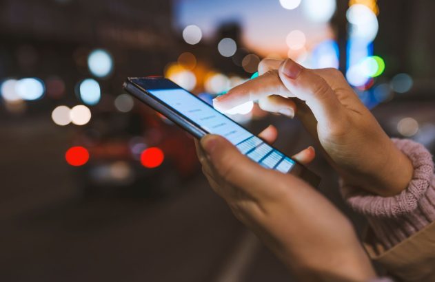 Mãos iluminadas usando smartphone no escuro, luzes borradas de cidade ao fundo.