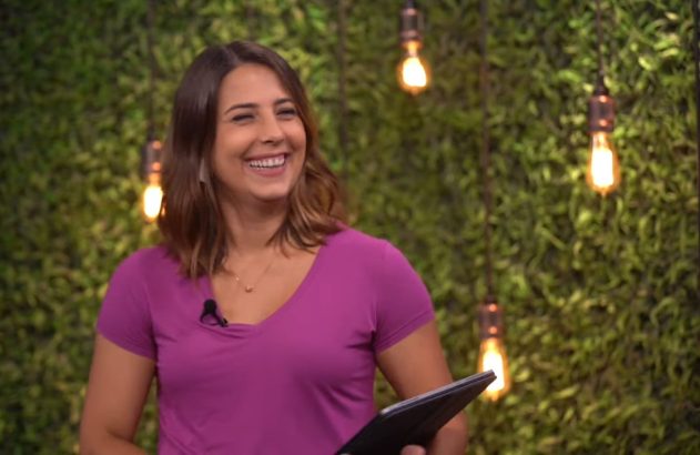 Mulher sorridente de camisa roxa segurando um tablet, com microfone. Fundo verde com lâmpadas penduradas.