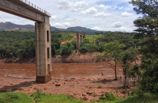 Viaduto de concreto sobre vale com rio e margens cobertas por lama marrom-avermelhada. Pessoas observam.