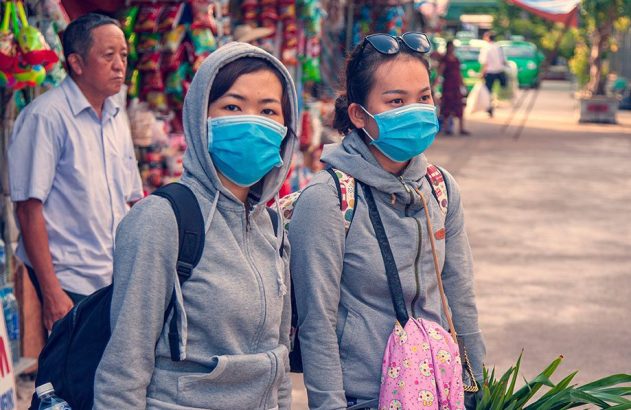 Duas mulheres usando máscaras azuis em uma rua. Um homem está em segundo plano.