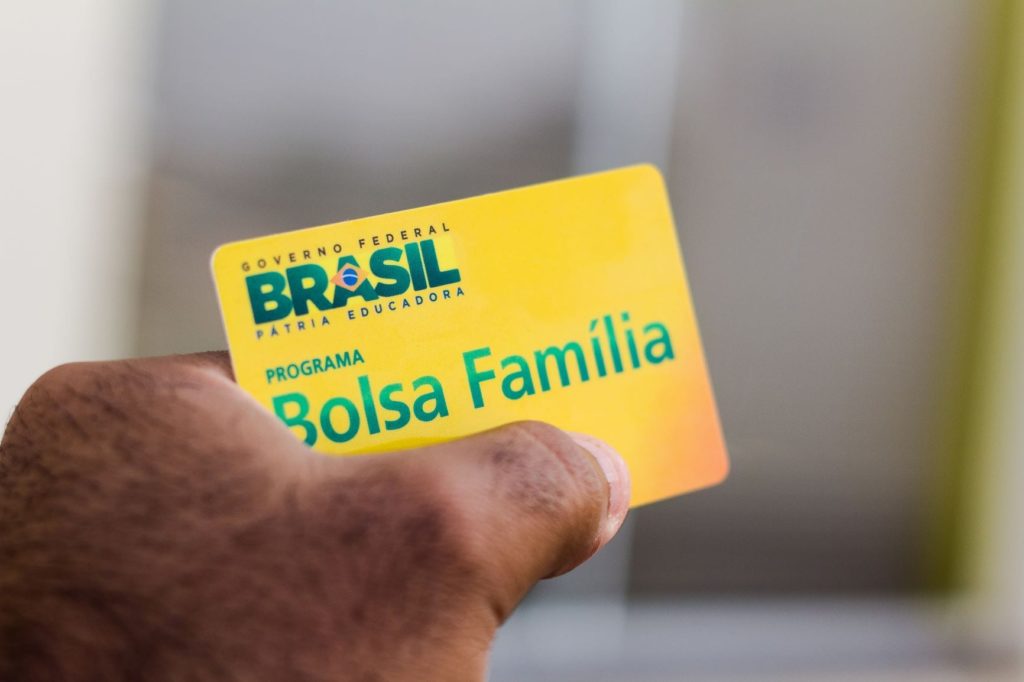Mão segura cartão amarelo do programa "Bolsa Família" com a marca "Brasil - Pátria Educadora".