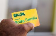 Mão segura cartão amarelo do programa "Bolsa Família" com a marca "Brasil - Pátria Educadora".