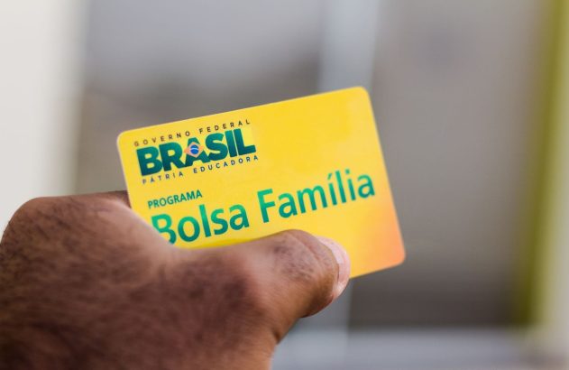 Mão segura cartão amarelo do programa "Bolsa Família" com a marca "Brasil - Pátria Educadora".