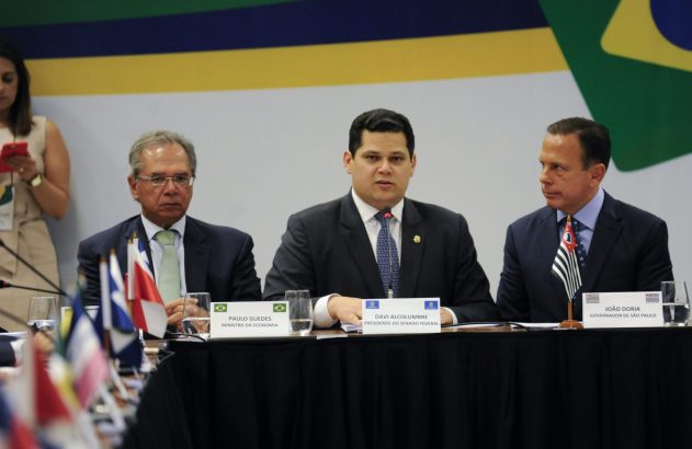 Três homens de terno sentados em mesa de reunião formal. Há bandeiras sobre a mesa e um grande banner ao fundo.