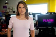 Mulher de camisa rosa posa em escritório. Monitores de computador visíveis no fundo.