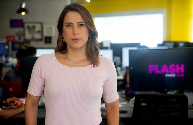 Mulher de camisa rosa posa em escritório. Monitores de computador visíveis no fundo.