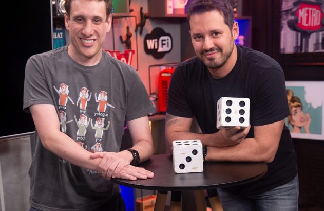 Dois homens sorrindo em estúdio. Um segura dado branco, e outro dado na mesa.