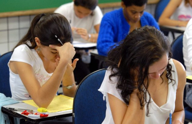Alunos em sala de aula, sentados em mesas, concentrados em papéis. Ambiente de estudo ou prova.