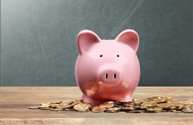 Um cofrinho de porquinho rosa rodeado por moedas douradas em uma mesa de madeira, com um quadro negro ao fundo.