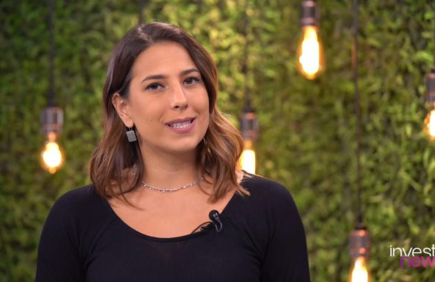 Apresentadora sorrindo em blusa preta e microfone, contra parede de musgo verde iluminada por lâmpadas. Logo Invest News.