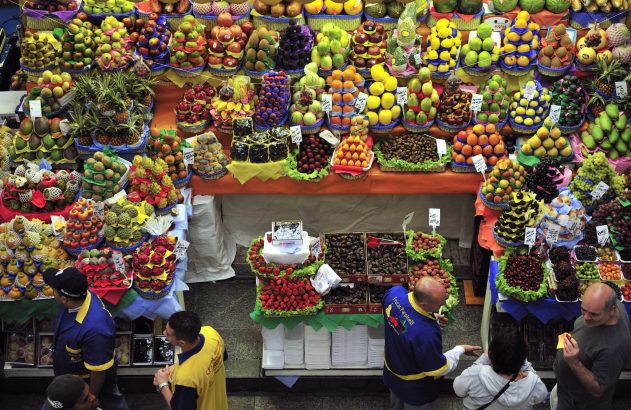 Vista aérea de um mercado de frutas com barracas repletas de pilhas coloridas de frutas e pessoas.