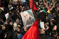 Multidão reunida, com retratos de Qasem Soleimani e bandeiras vermelhas e pretas.