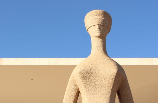 Escultura humana estilizada e bege com faixa nos olhos, sob céu azul e parede clara.