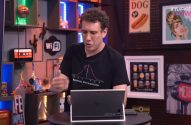 Homem de camiseta escura gesticulando e apresentando, com notebook à frente, em um estúdio.