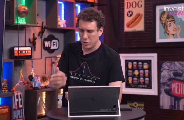 Homem de camiseta escura gesticulando e apresentando, com notebook à frente, em um estúdio.