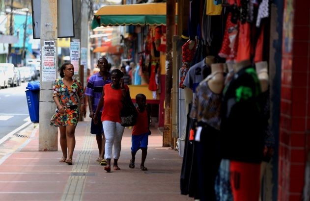 Mulheres e criança caminham em calçada movimentada com lojas e roupas à venda.