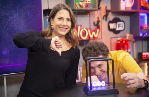 Mulher sorrindo faz "não gostei"; homem sorrindo brinca com pêndulo de Newton iluminado.