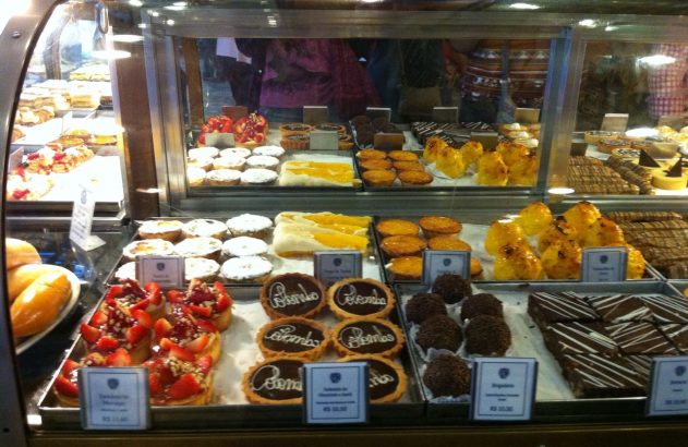 Vitrine de confeitaria com doces diversos: tortas de morango, chocolate, brigadeiros e quindins.