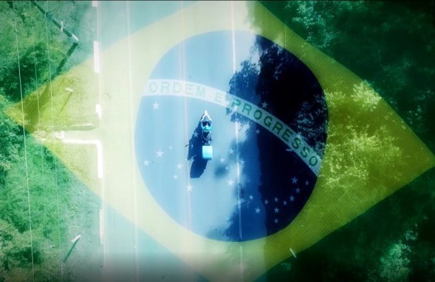 Motoqueiro com caixa de entrega em estrada, visto de cima. Bandeira do Brasil sobreposta transparentemente à cena verde.