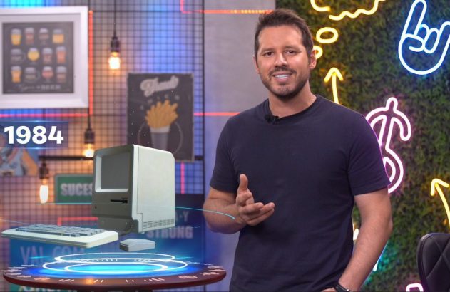 Homem sorrindo apresenta um computador Macintosh de 1984 projetado em um cenário de estúdio.