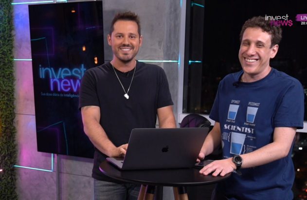 Dois apresentadores sorrindo em estúdio com laptop e tela "invest news" ao fundo.
