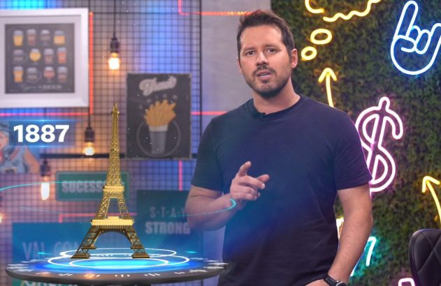 Homem em estúdio moderno, apontando. Miniatura da Torre Eiffel em mesa, luzes neon no fundo.