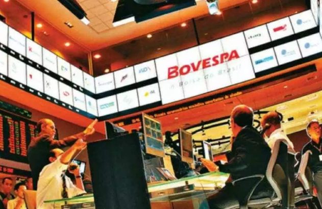 Corretores negociam ativamente em pregão da bolsa BOVESPA, com telas e painéis de dados ao fundo.