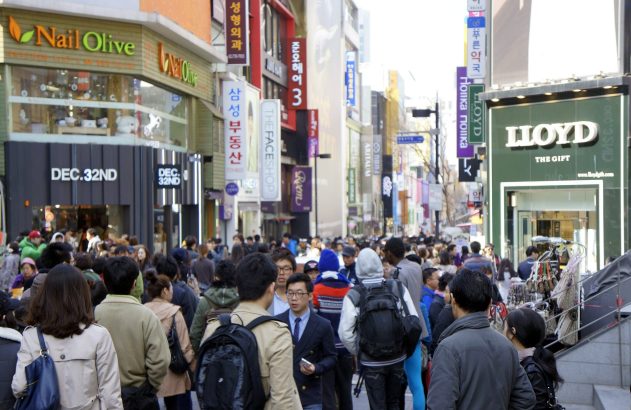 Pessoas caminham em rua comercial movimentada, ladeada por lojas e edifícios.