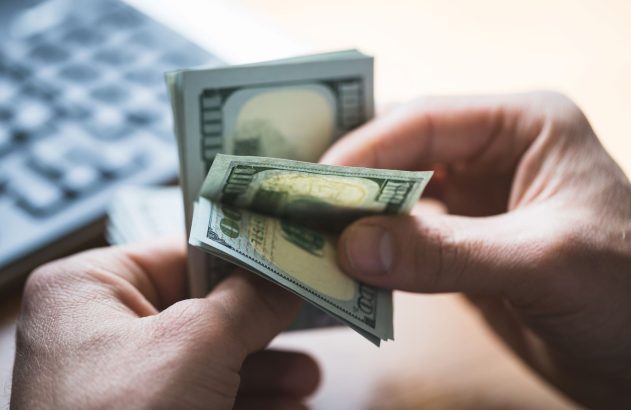 Mãos contam um maço de notas de cem dólares, com teclado desfocado no fundo.