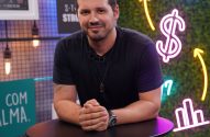 Homem sorrindo, de camiseta escura, apoiado em mesa. Fundo com luzes neon financeiras.