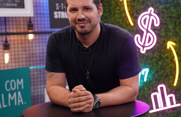Homem sorrindo, de camiseta escura, apoiado em mesa. Fundo com luzes neon financeiras.