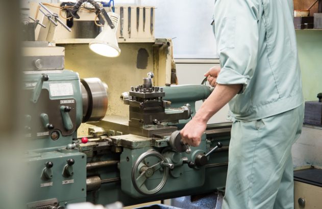 Homem em uniforme claro operando um torno mecânico em ambiente industrial.