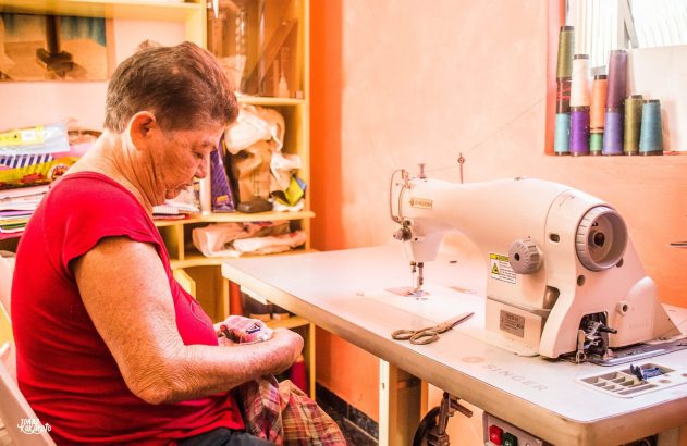 Mulher sentada manuseando tecido, com máquina de costura Singer e carretéis de linha no fundo.