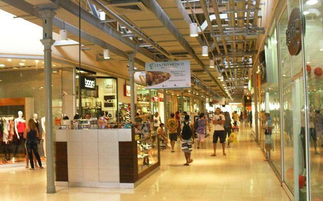 Interior de shopping com pessoas caminhando entre lojas e quiosque.