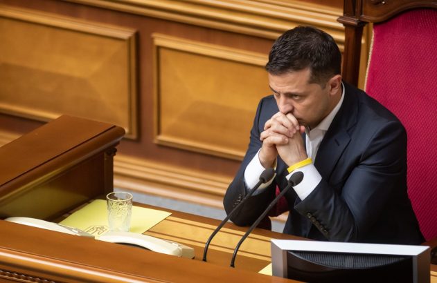 Homem de terno com pulseira azul e amarela, sentado à mesa com microfones, mãos unidas. Ambiente formal.