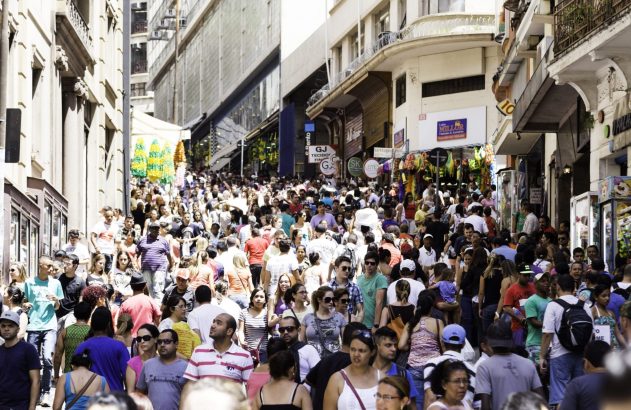 Multidão de pessoas caminhando em uma rua íngreme e movimentada, ladeada por lojas e edifícios urbanos.