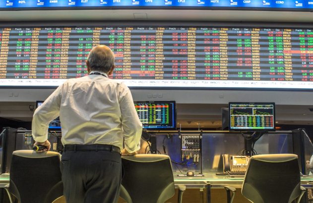 Homem de costas observa um grande painel digital com dados da bolsa de valores em uma sala de operações.