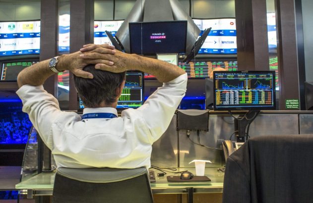 Operador financeiro de costas, com as mãos na cabeça, olhando gráficos e dados em múltiplas telas.