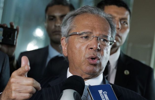 Homem de terno e óculos fala em microfones, apontando com a mão. Outras pessoas desfocadas no fundo.