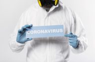 Pessoa com traje de proteção branco, máscara e luvas azuis, segurando placa "CORONAVIRUS" e apontando.