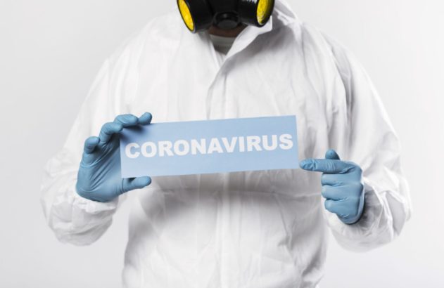 Pessoa com traje de proteção branco, máscara e luvas azuis, segurando placa "CORONAVIRUS" e apontando.