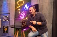 Homem barbudo sorrindo aponta para laptop sobre mesa, com luzes neon de dólar e ideia.