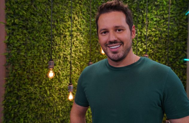 Homem sorrindo de camiseta verde em frente a uma parede de folhagem com luzes.