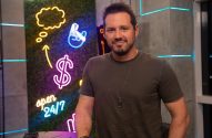 Homem sorrindo. Fundo com painel de folhagem verde e luzes neon: símbolo de dólar, balão de fala e 'open 24/7'.