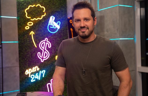 Homem sorrindo. Fundo com painel de folhagem verde e luzes neon: símbolo de dólar, balão de fala e 'open 24/7'.