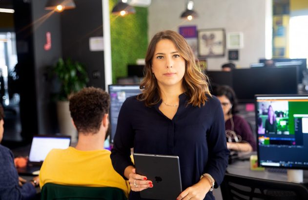 Mulher segura tablet em escritório, pessoas trabalhando ao fundo.