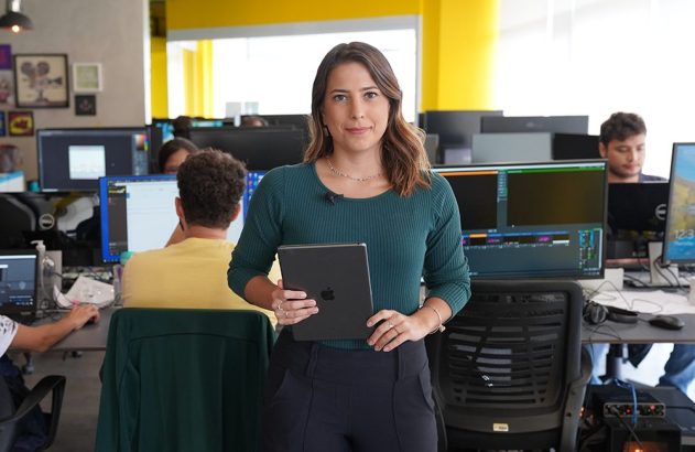 Mulher em pé, segurando um tablet, em um escritório com colegas trabalhando ao fundo.