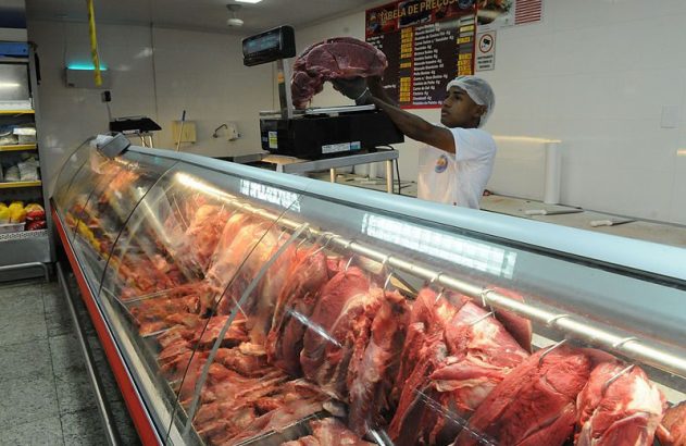 Funcionário de açougue pesa carne em balança, com grande balcão refrigerado de carnes vermelhas em exposição.